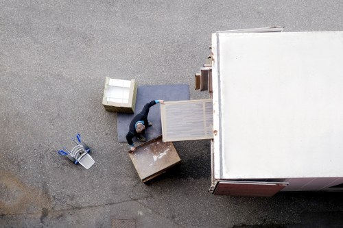 An overhead view of a man in a dark jacket and blue jeans standing on a residential street driveway, surrounded by various household items including a small white storage chest, a grey cushion, a wooden cabinet with a light finish, a small brown box, and a cardboard box, all arranged on the grey tarmac surface. The man appears to be in the process of loading or organizing furniture and belongings, with a large white moving van parked adjacent to the driveway, indicating a house removal scenario. Nearby, a pair of work gloves and a metal trolley are also visible, suggesting ongoing packing or unloading activity typical of professional house removals in the Barnes, London area. The scene is lit with natural daylight, capturing a clear view of the furniture and personal items ready for transport, with residential buildings in the background, aligning with the services offered by Barnes Man with Van in the local postcode district.