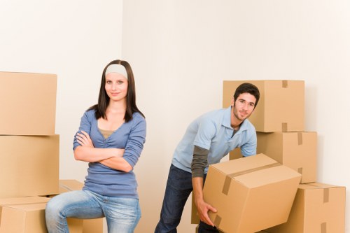 A young man and woman are in a bright, neutral-toned room, surrounded by stacked cardboard moving boxes of varying sizes, some sealed with packing tape. The woman, with shoulder-length dark hair, is seated on one of the boxes with her arms crossed and a neutral expression, wearing a blue long-sleeved top and light blue jeans. The man, with short dark hair and a slight smile, is standing next to him, leaning slightly forward while holding a large, sealed cardboard box in both hands. The background features a plain off-white wall, suggesting a house interior in a residential area such as Barnes or nearby postcode areas, consistent with local removals services. The scene captures a moment during a house move, with no visible furniture or appliances, emphasizing the focus on packing and relocation. The natural lighting highlights the textures and colors of the cardboard boxes, clothing, and minimalistic indoor environment, illustrating professional house removal activity typical of Barnes Man with Van services.